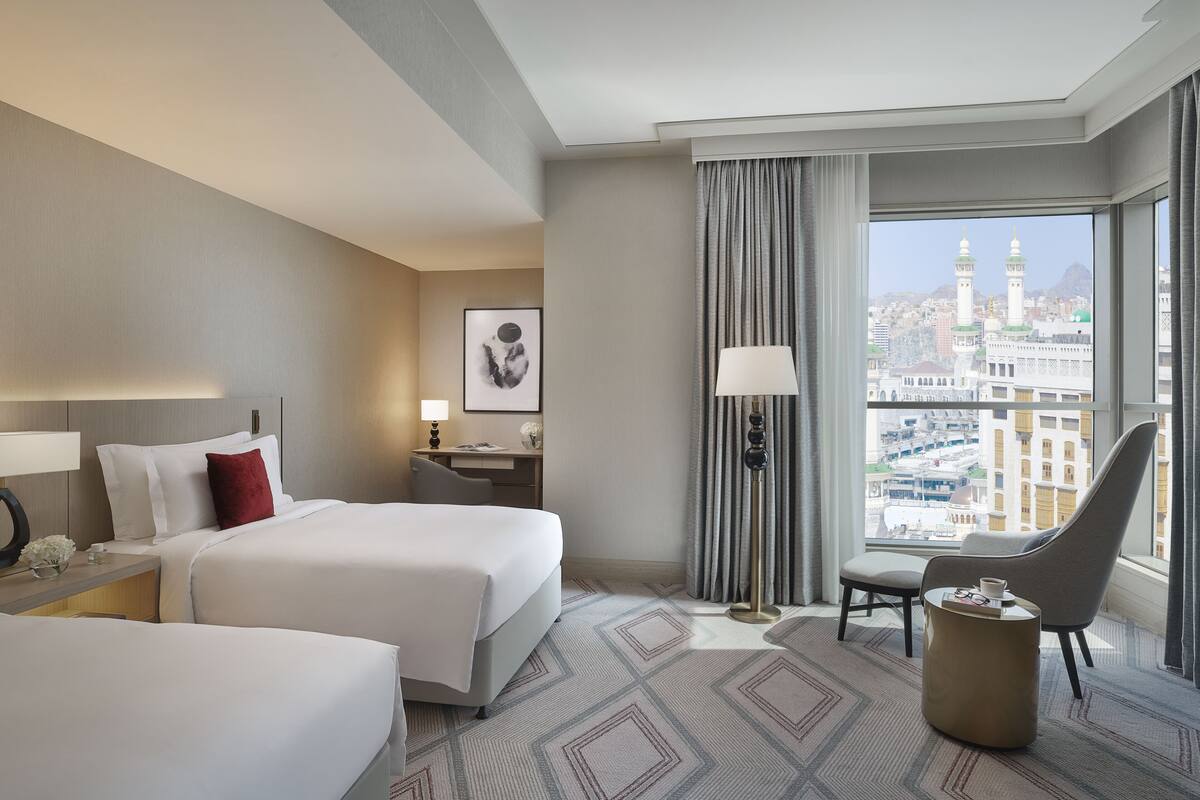 A hotel room with two beds, a desk, a chair, a lamp, and a view of a cityscape with a mosque.