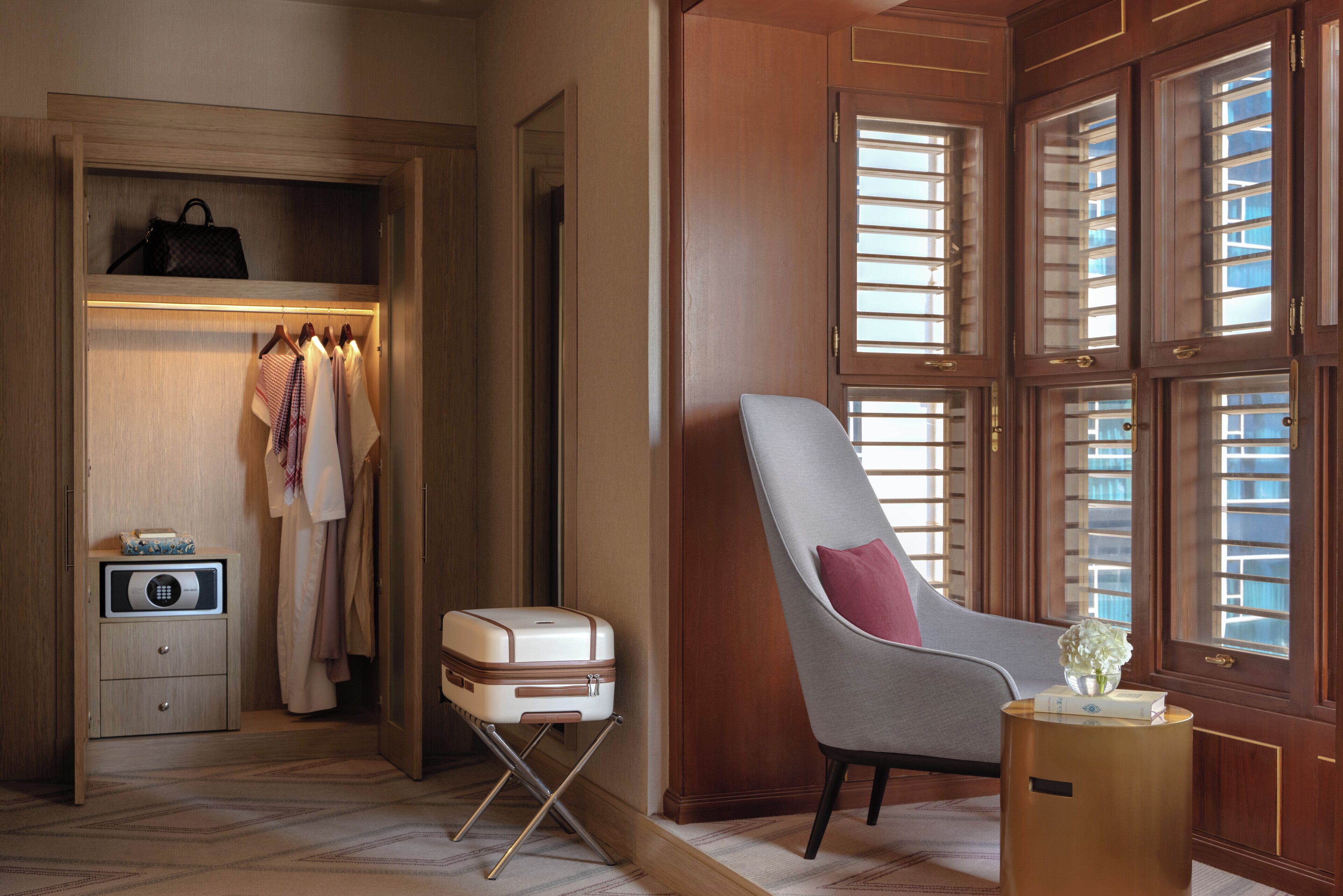 Une pièce comprenant une armoire en bois, un fauteuil gris, une petite table de chevet et une fenêtre avec des stores.