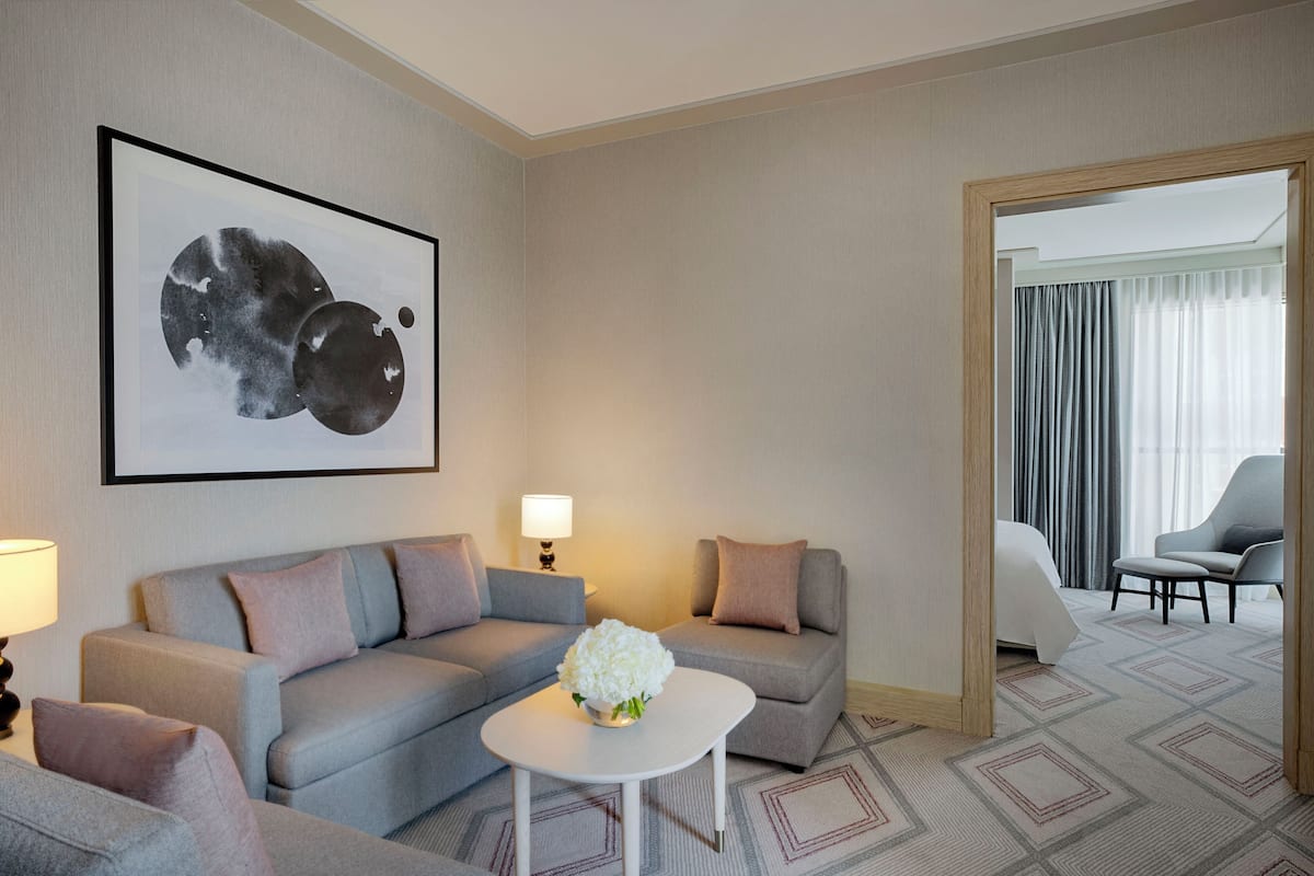 A hotel room with a grey sofa set, a small white table with a vase of flowers, a framed abstract painting, and a view of a balcony with a grey chair.