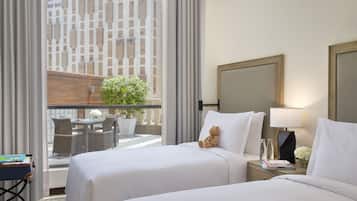 A hotel room with two beds, a large window with curtains, a potted plant, a table with chairs, and a view of a building outside.