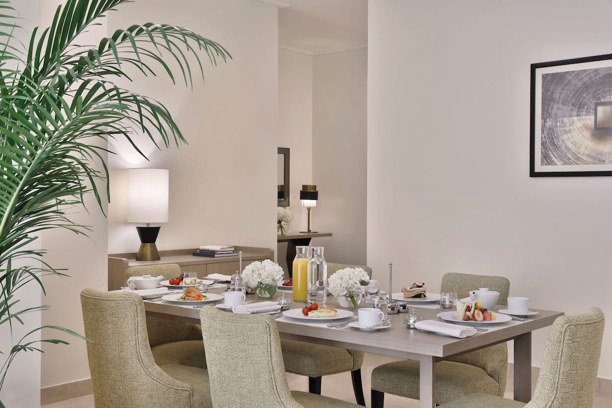 A dining area with a long table set for four, surrounded by upholstered chairs. There are floral centerpieces on the table, a lamp, and a framed artwork on the wall.