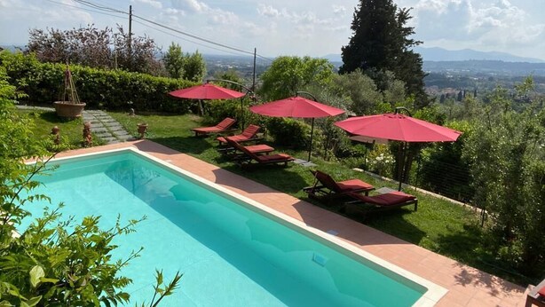 Una piscina con sombrillas rojas y sillas de descanso, rodeada de vegetación y con vistas a montañas lejanas.