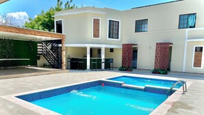Una piscina con borde de baldosas, un área de estar techada y un edificio de dos plantas con ventanas y una puerta.
