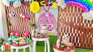 Un espacio interior decorado con una valla de madera, una maqueta de una casa rosa, y mesas con cupcakes y pequeñas plantas.
