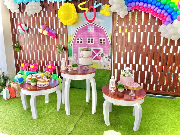 A decorated indoor space with a wooden fence, a pink house model, and tables with cupcakes and small plants.