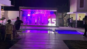 Un área de piscina al aire libre con un escenario decorado con globos y un letrero con el número '15', rodeado por un edificio moderno con una escalera y una zona de estar.