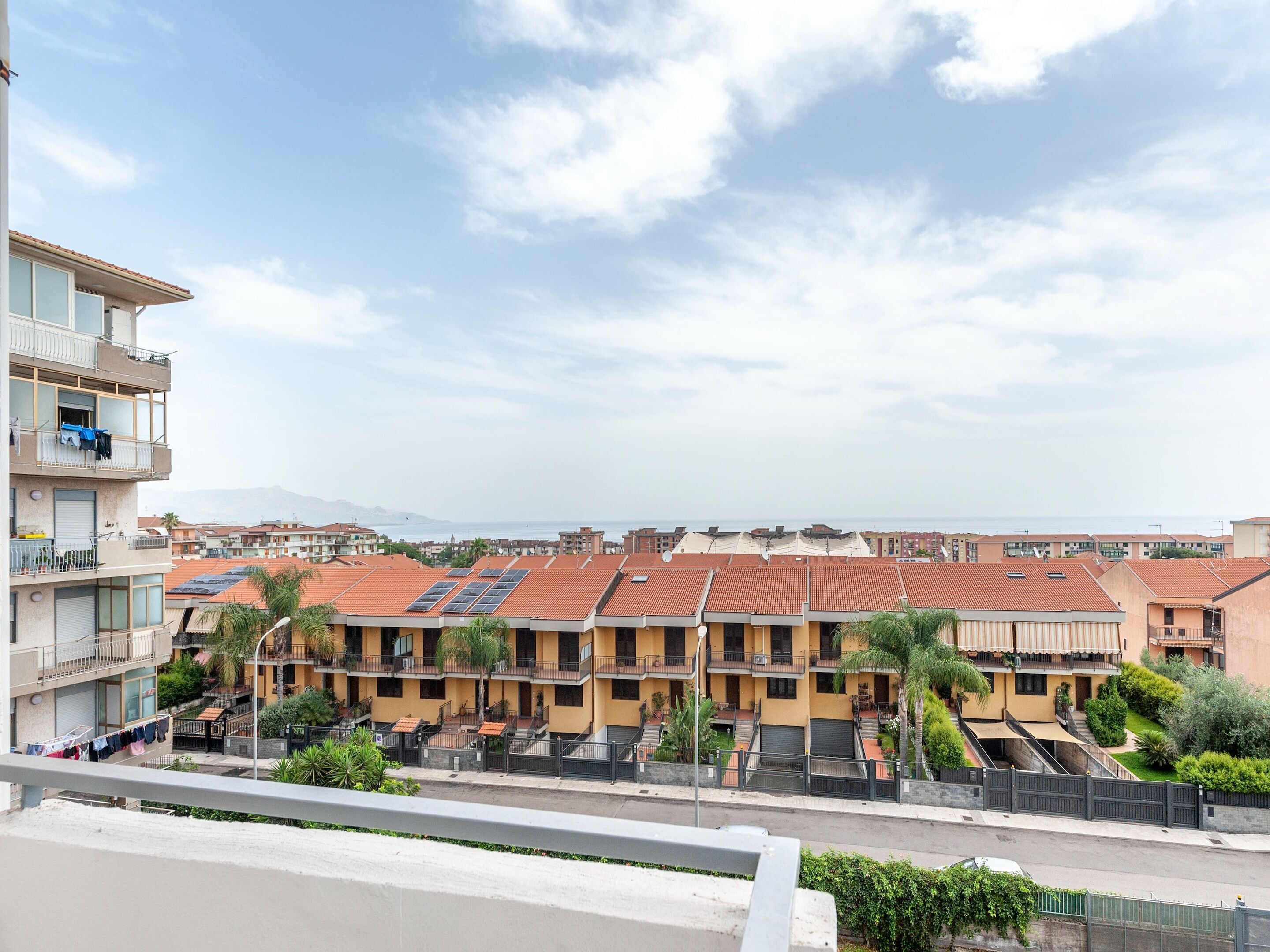 A residential building with multiple balconies, solar panels on the roof, and a view of the sea in the background.