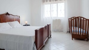 Un dormitorio con cama de madera, una mesita de noche con lámpara, una cuna y una ventana con cortinas traslúcidas.