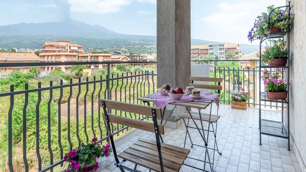Un balcón con una mesa y sillas, una planta en maceta y vistas a edificios y montañas al fondo.
