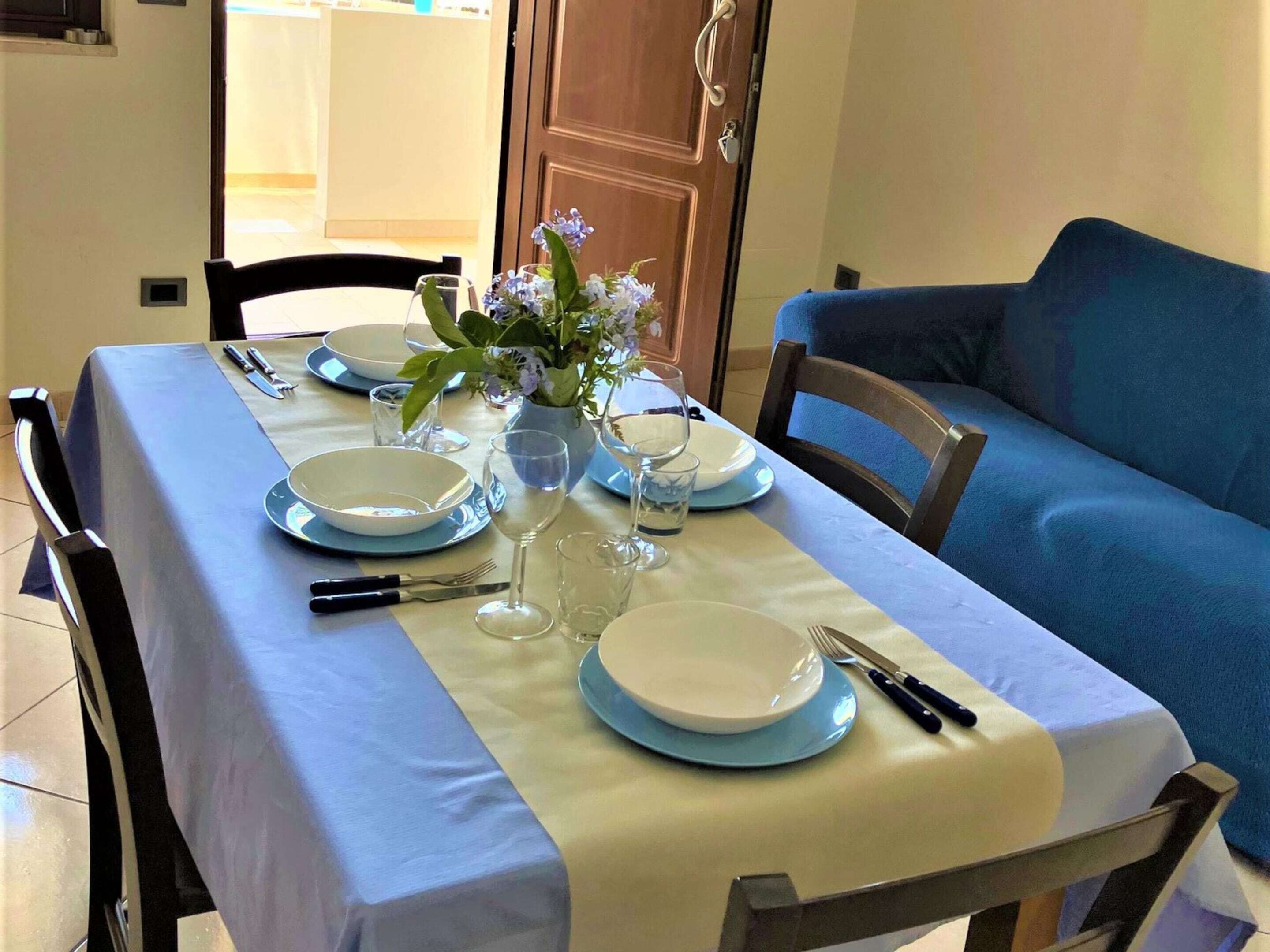 A dining table set with plates, glasses, and cutlery, a vase with flowers, and a blue sofa in the background.