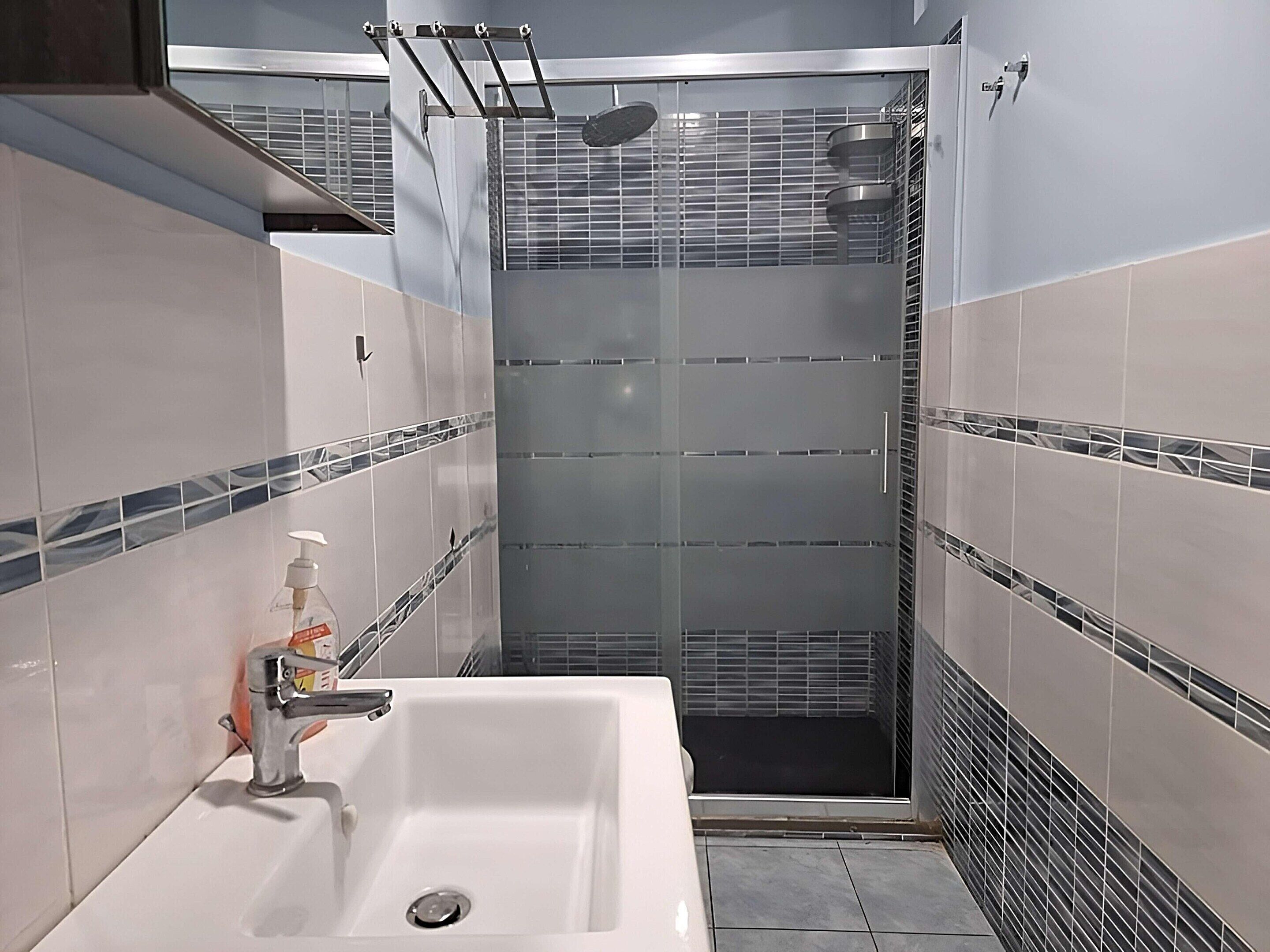 Ein modernes Badezimmer mit einer ein Glasfach raumtrennenden Duschkabine, einem weißen Waschbecken mit Seifenspender und Fliesenwänden mit blau-weißem Muster.