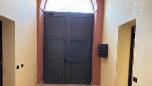 Een gang met een grote, donkere deur aan het einde, een klein intercomsysteem op de muur en een vloer met een patroon.