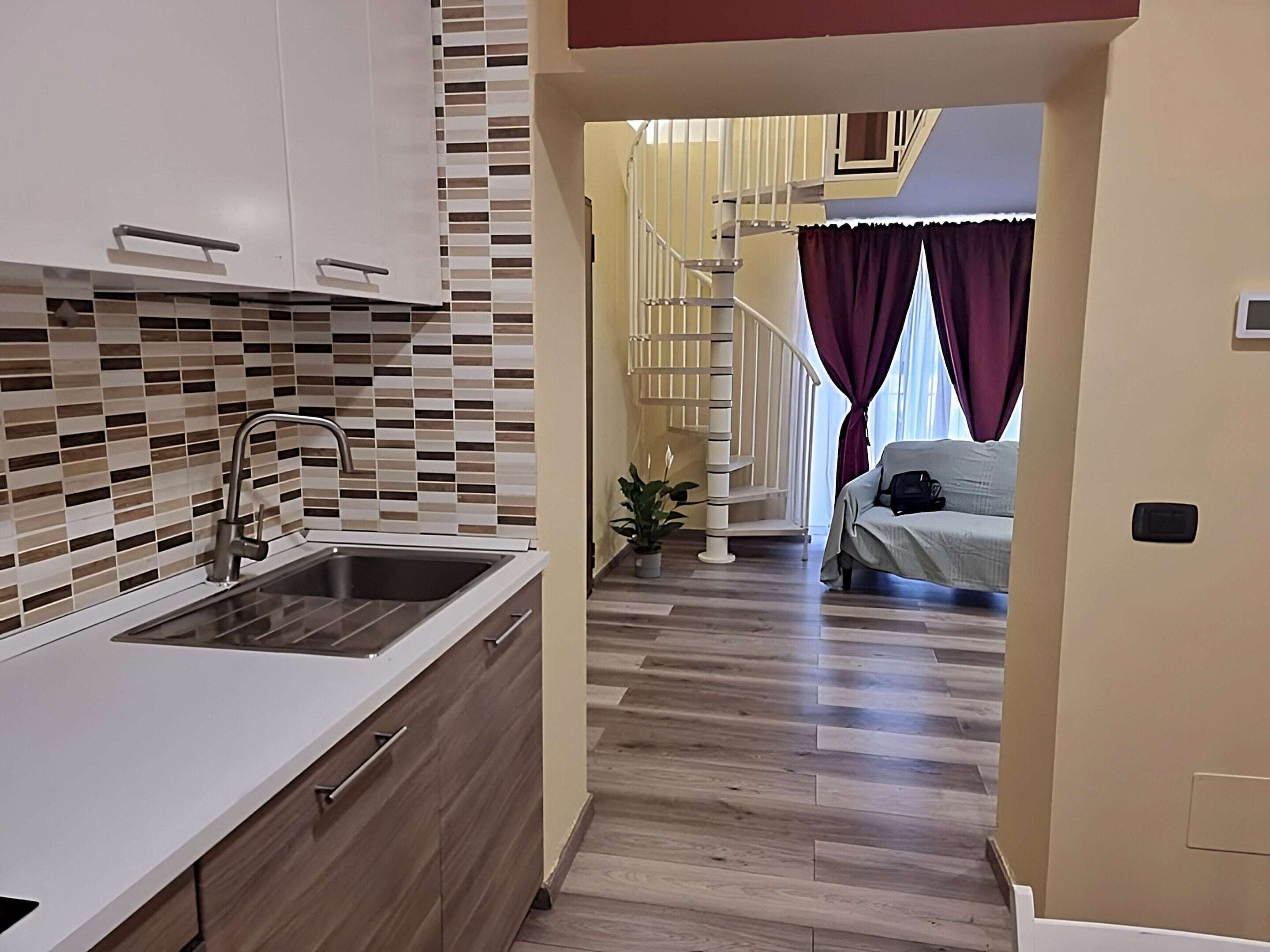 A modern kitchen with a sink and cabinets, a hallway with wooden flooring, a staircase, and a bed in the background.