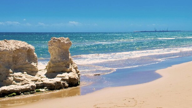 Ein Strand mit klarem, blauem Wasser, sandigem Ufer und Felsformationen.