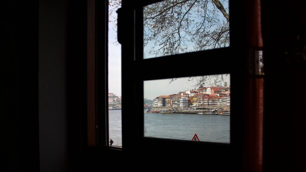 A view through a window with a view of a waterfront cityscape, including buildings and a body of water. - Estúdio vista rio Douro (Vila Nova de Gaia)