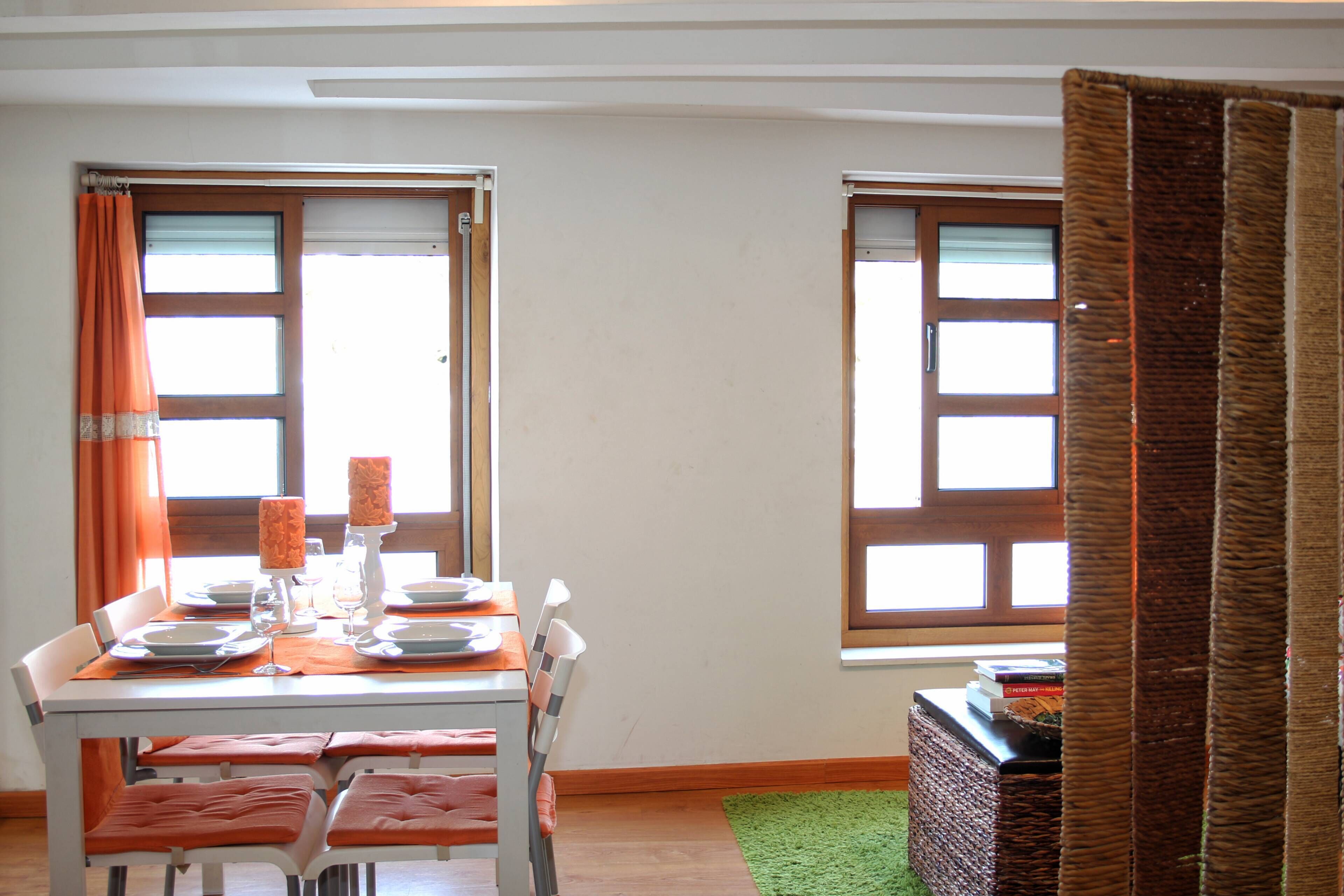 A dining area with a table set for four, two windows with curtains, and a green rug.