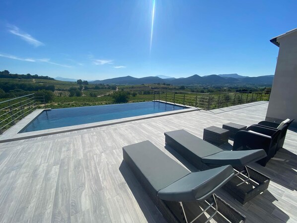A rooftop infinity pool with a view of vineyards and mountains, outdoor seating, and a clear sky.