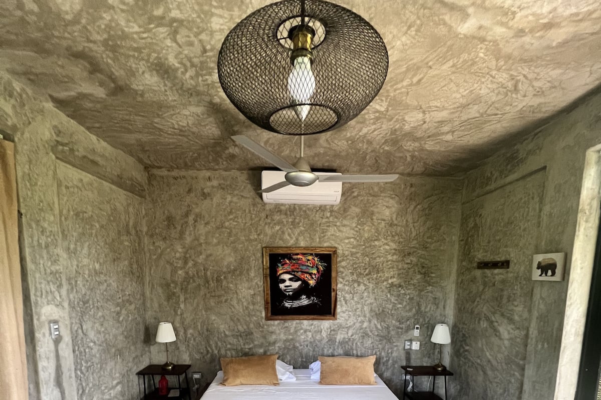 A room with a bed, two bedside tables, a ceiling fan, a wall-mounted air conditioner, and a framed picture on the wall.