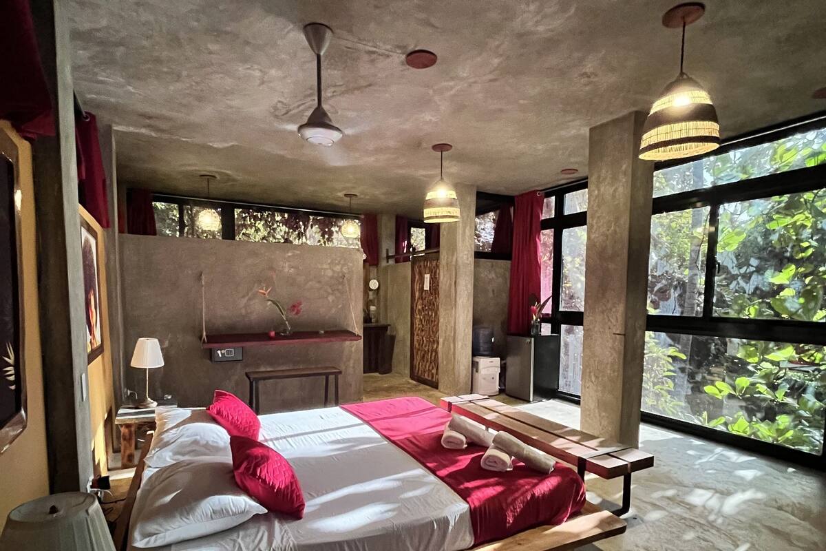 A bedroom with a large bed, wooden headboard, two pendant lights, a window with a view of greenery, and a ceiling fan.
