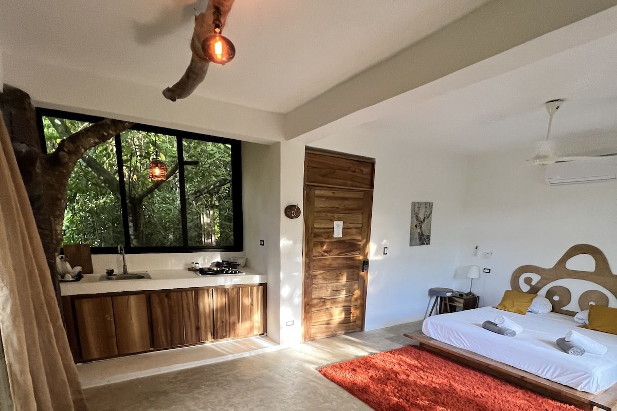 A bedroom with a large bed, a red rug, a grey sofa, a wooden door, and a window with a view of trees.