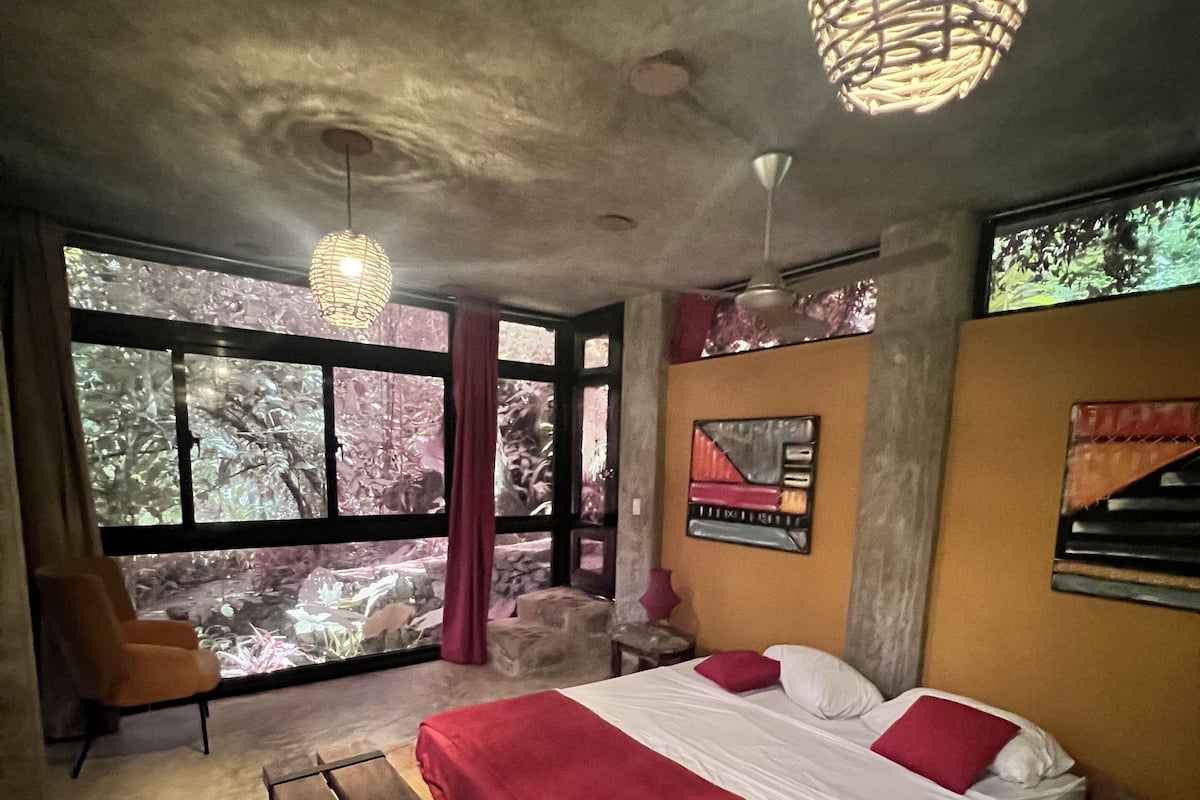 A bedroom with a large bed, a bench, a chair, and a view of trees through large windows.