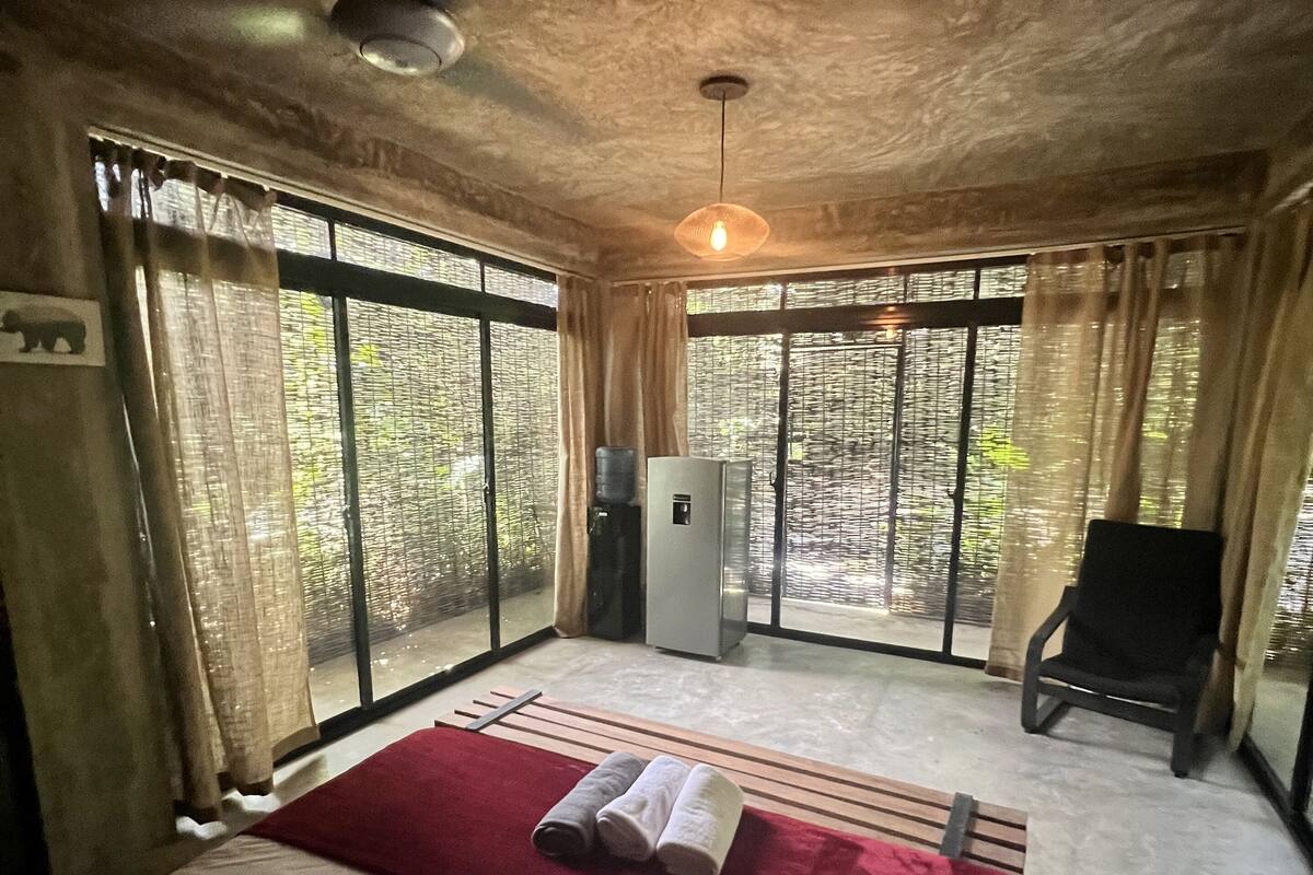 A bedroom with a bed, a red blanket, a ceiling fan, and a view of trees through large windows.