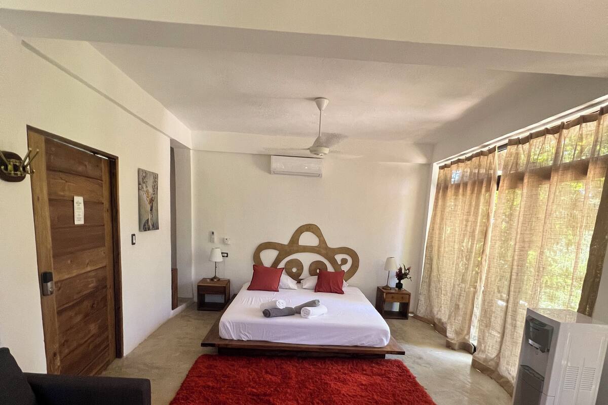 A bedroom with a bed, a red rug, a ceiling fan, a wooden door, and a window with curtains.