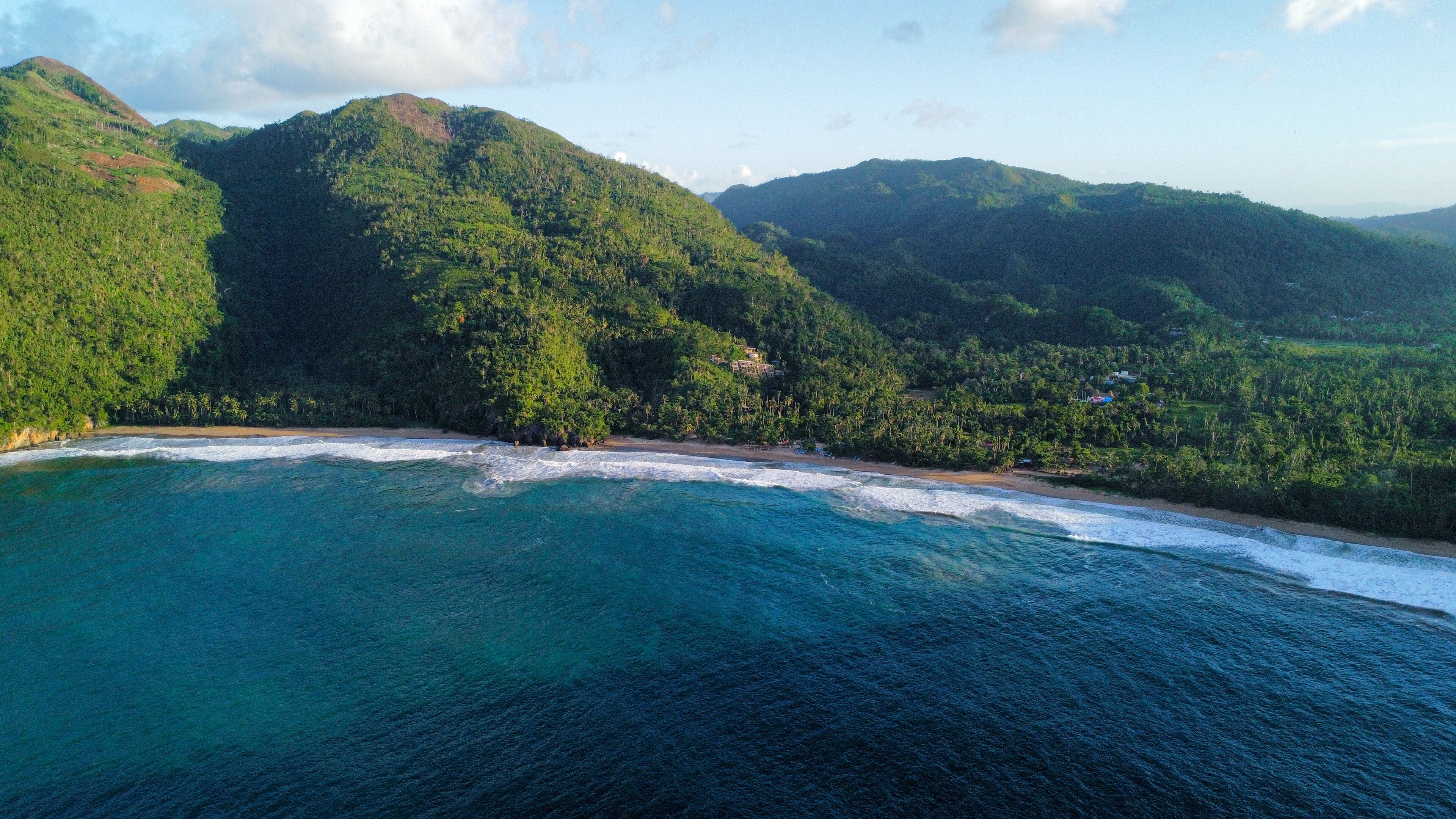 A coastal landscape with a beach, forested hills, and clear blue waters.