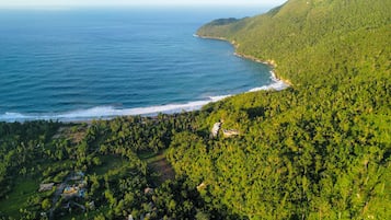 Vue aérienne d’une zone côtière caractérisée par une végétation luxuriante, une plage et un petit complexe touristique avec des bâtiments et des allées bien visibles.