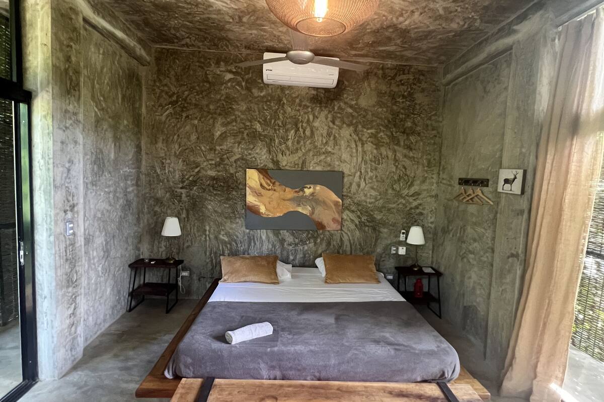 A bedroom with a bed, a ceiling fan, a wall-mounted air conditioner, a painting, two bedside tables with lamps, and a window with curtains.