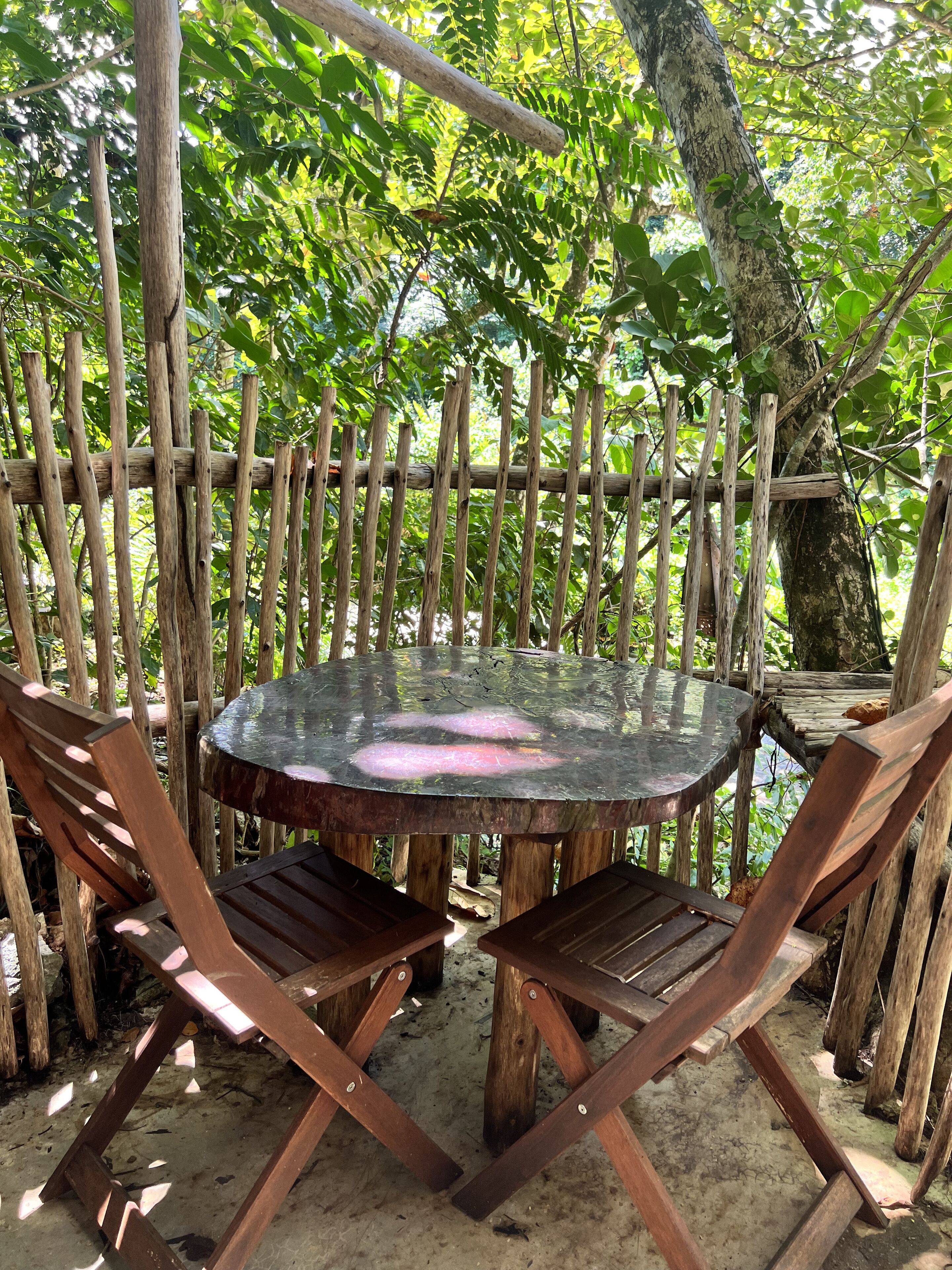 A round wooden table with metal legs, surrounded by wooden chairs, set against a backdrop of a wooden fence and lush greenery.