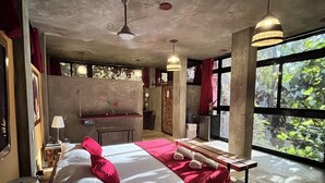 A bedroom with a large bed, a wooden bench, a desk, and a view of greenery through large windows. - Taino Beach Lofts (Samaná)