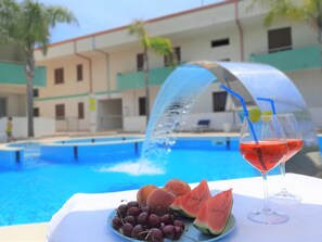 A poolside table with a plate of fruit, a glass of red drink, and a water fountain. - Belvilla by OYO Appartamento Lato Piscina (Ispica)