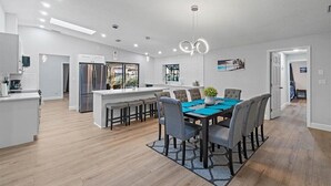 A modern kitchen with a central island, a dining area with a blue tablecloth, and a view of the outdoors through a large window. - Heated Saltwater Pool & Spa l Family Friendly l Near Beaches & Clearwater (Palm Harbor)