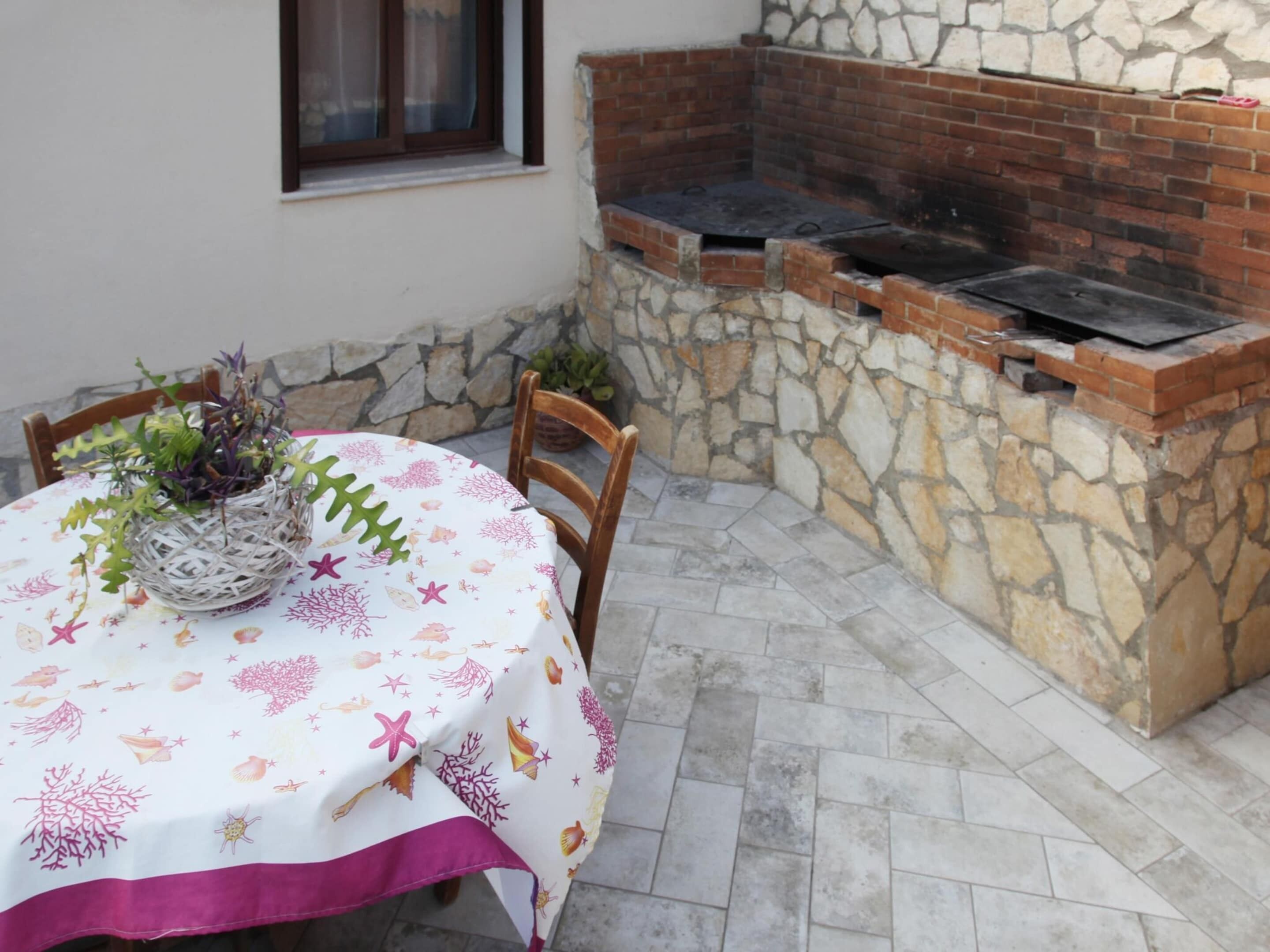 Une terrasse avec un mur en pierre, un four en briques, une table recouverte d’une nappe blanche et des chaises.