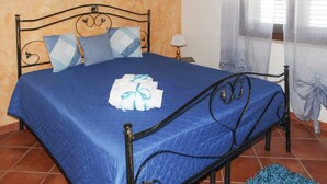 Una cama con colcha azul, estructura metálica y almohadas decorativas. Junto a la cama hay una mesita con una lámpara.