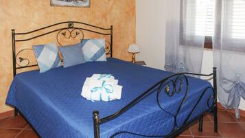 Cama com colcha azul, estrutura de metal e almofadas decorativas. Há uma pequena mesa com abajur ao lado da cama.