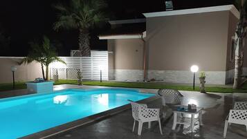 Piscina com deck revestido, cercada por mobiliário de jardim branco e palmeiras.