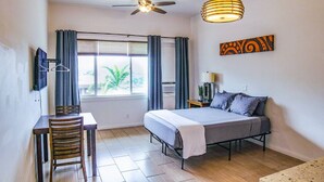 Un dormitorio con cama, escritorio, silla, ventilador de techo, ventana con cortinas y televisor montado en la pared.