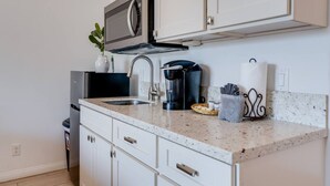 Una cocina con armarios blancos, microondas de acero inoxidable, cafetera y lavabo.