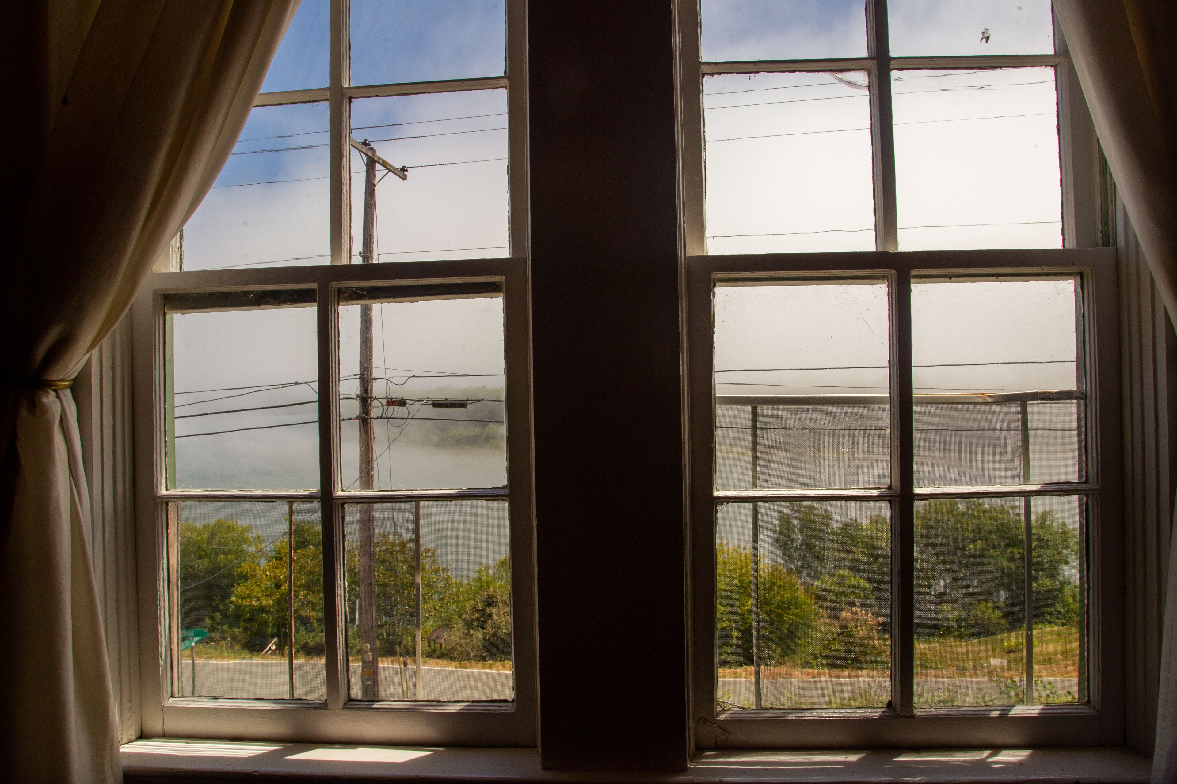 A window with a view of a road and trees, curtains partially drawn, and power lines visible outside.