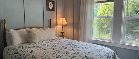 A bedroom with a bed, a nightstand with a lamp, a framed picture on the wall, and a window with curtains.