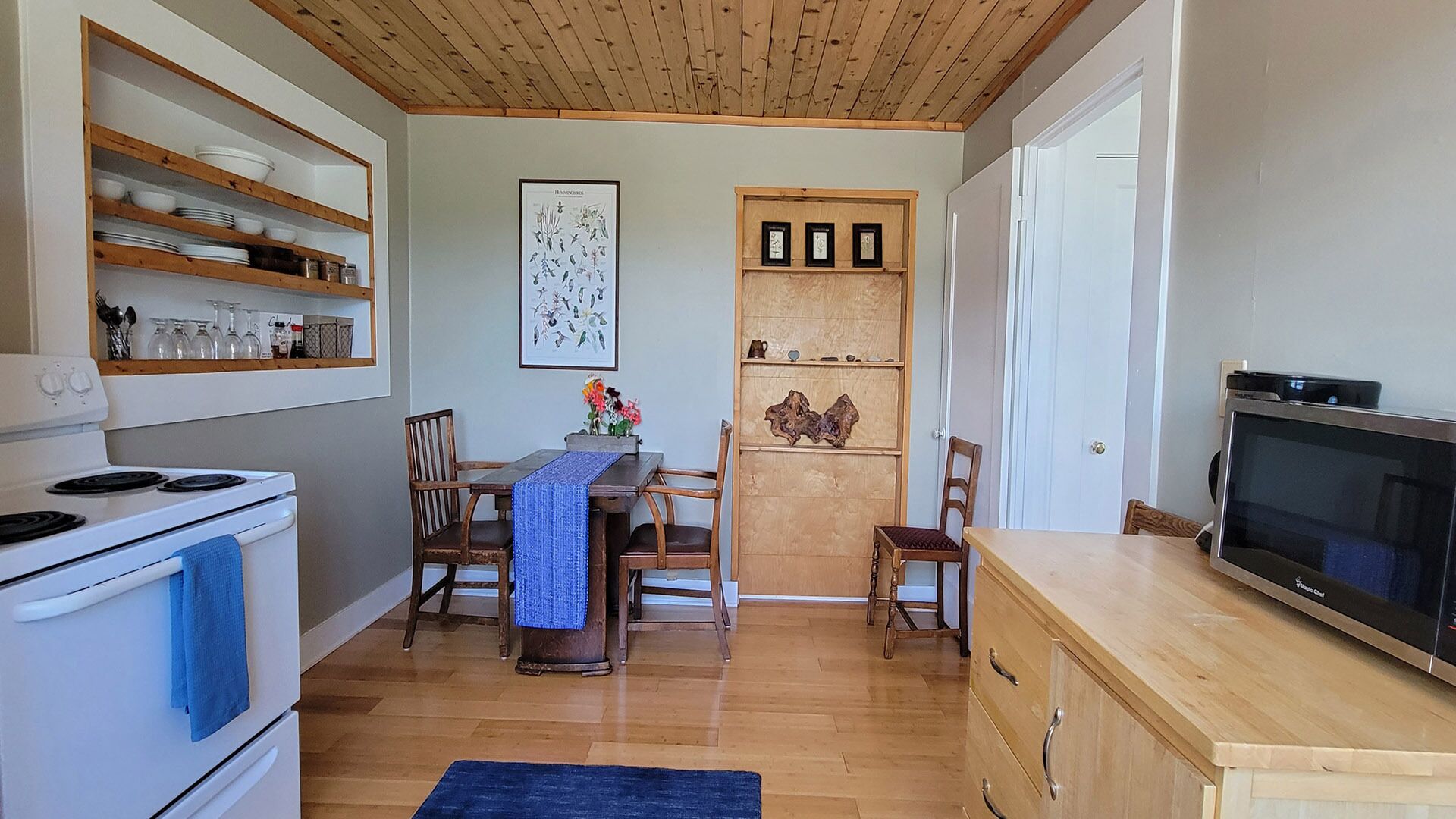 A compact kitchen with a stove, microwave, and wooden cabinets. There is a dining table with chairs and a blue rug.