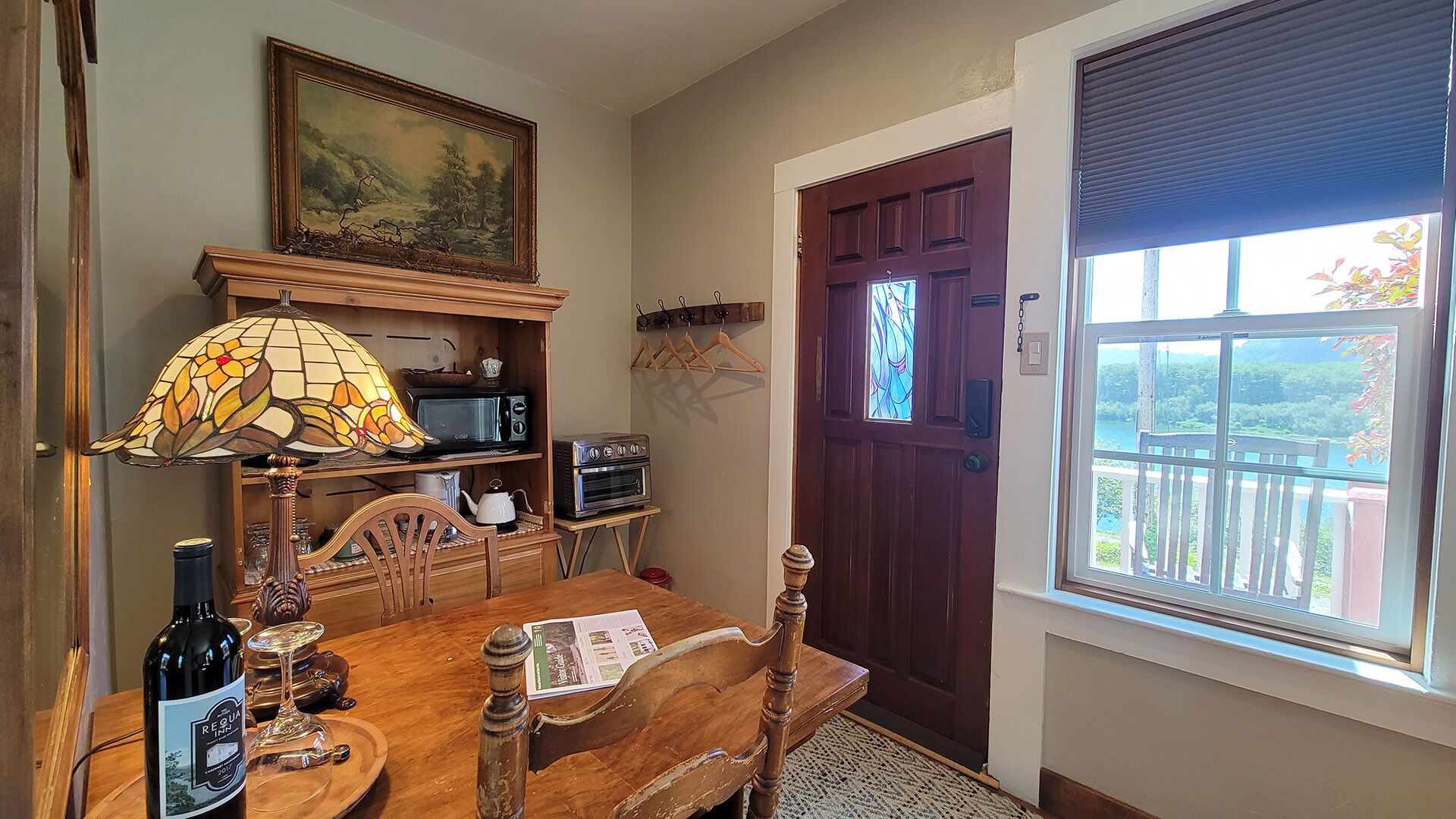 A dining area with a wooden table, chairs, a stained glass lamp, a microwave, a toaster, and a wine bottle.
