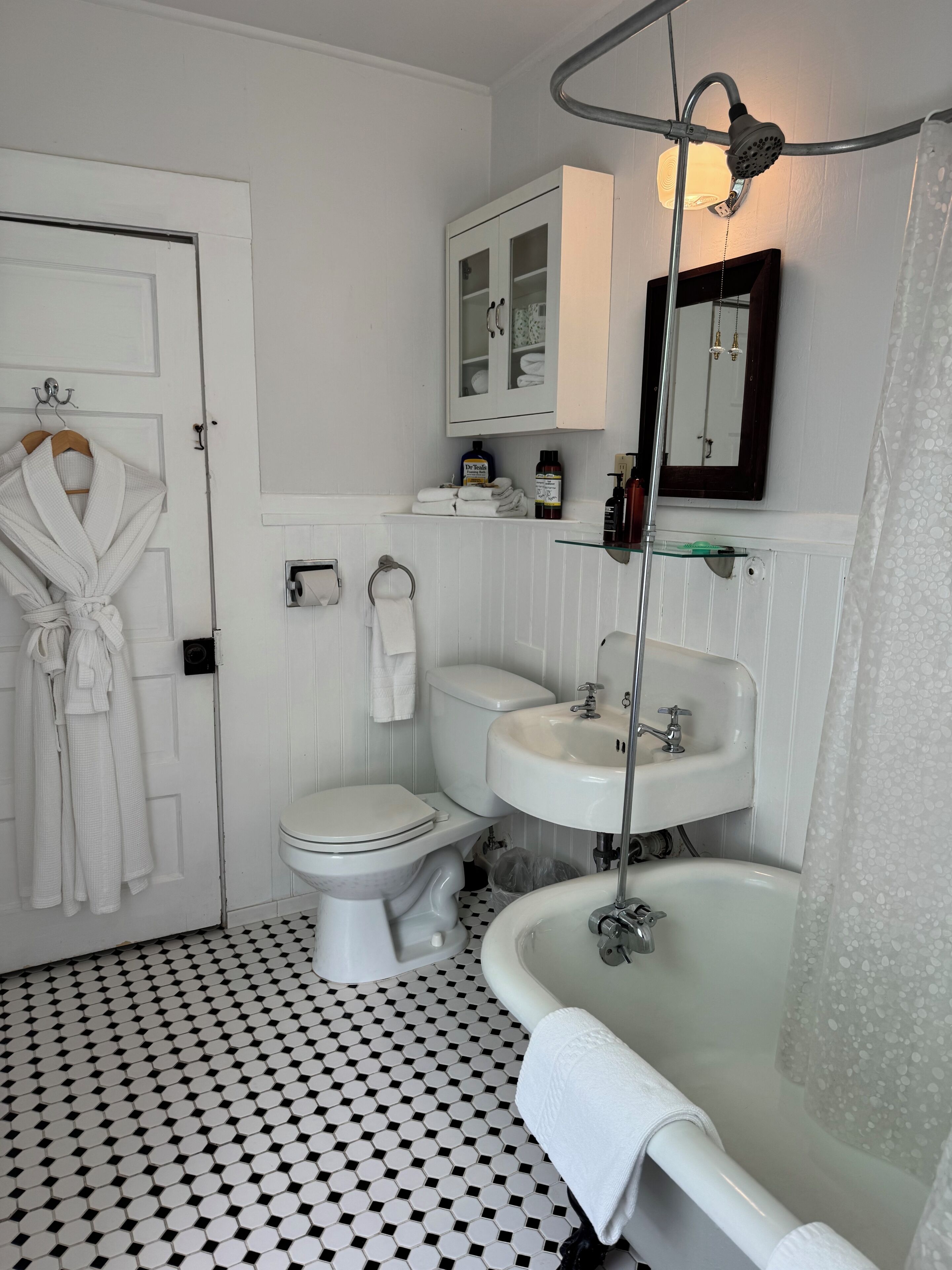 Une salle de bain équipée de toilettes, d’un lavabo et d’une baignoire. Une robe est accrochée à la porte, et des articles de toilette sont disposés sur le comptoir.