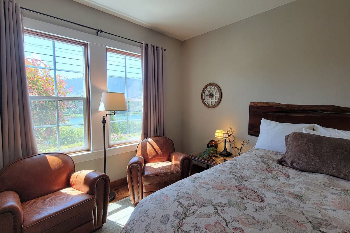 A hotel room with a large bed, two leather armchairs, a nightstand with a lamp, and a window with curtains.