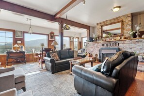 A spacious living room with a fireplace, leather sofas, a chess table, and a large window overlooking a mountainous landscape.