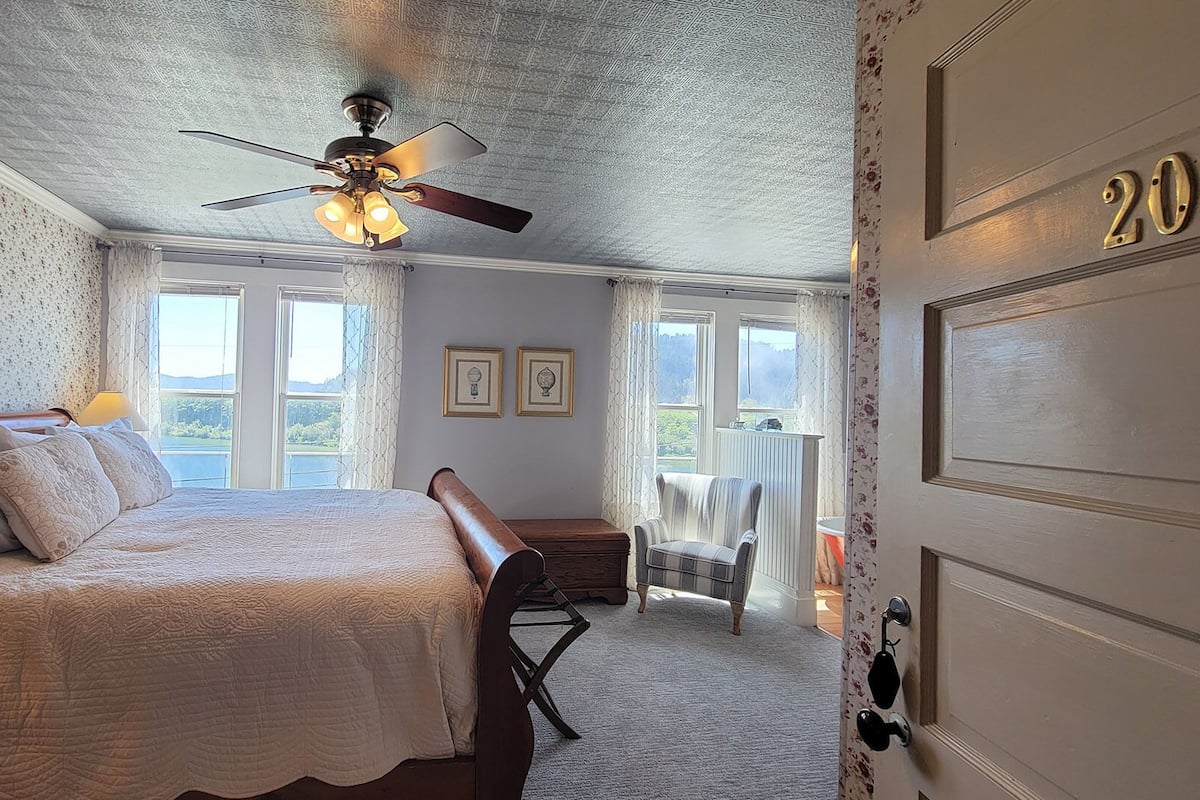 A bedroom with a bed, a ceiling fan, a bench, a chair, and a door with a number.