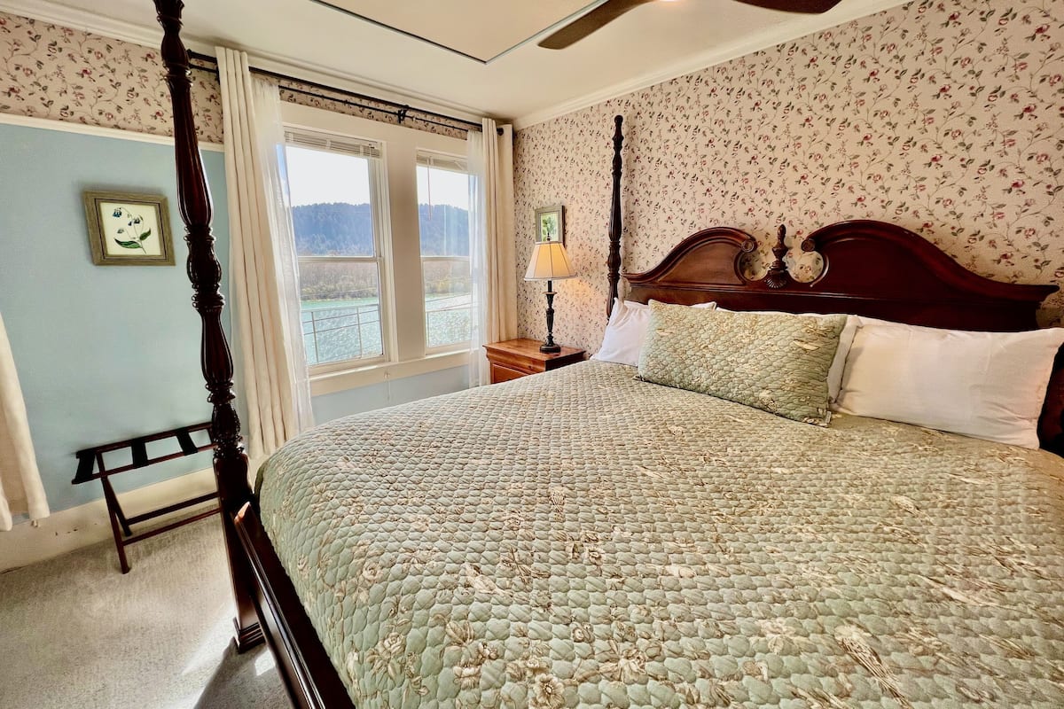A bedroom with a four-poster bed, floral wallpaper, and a view of a river through the window.