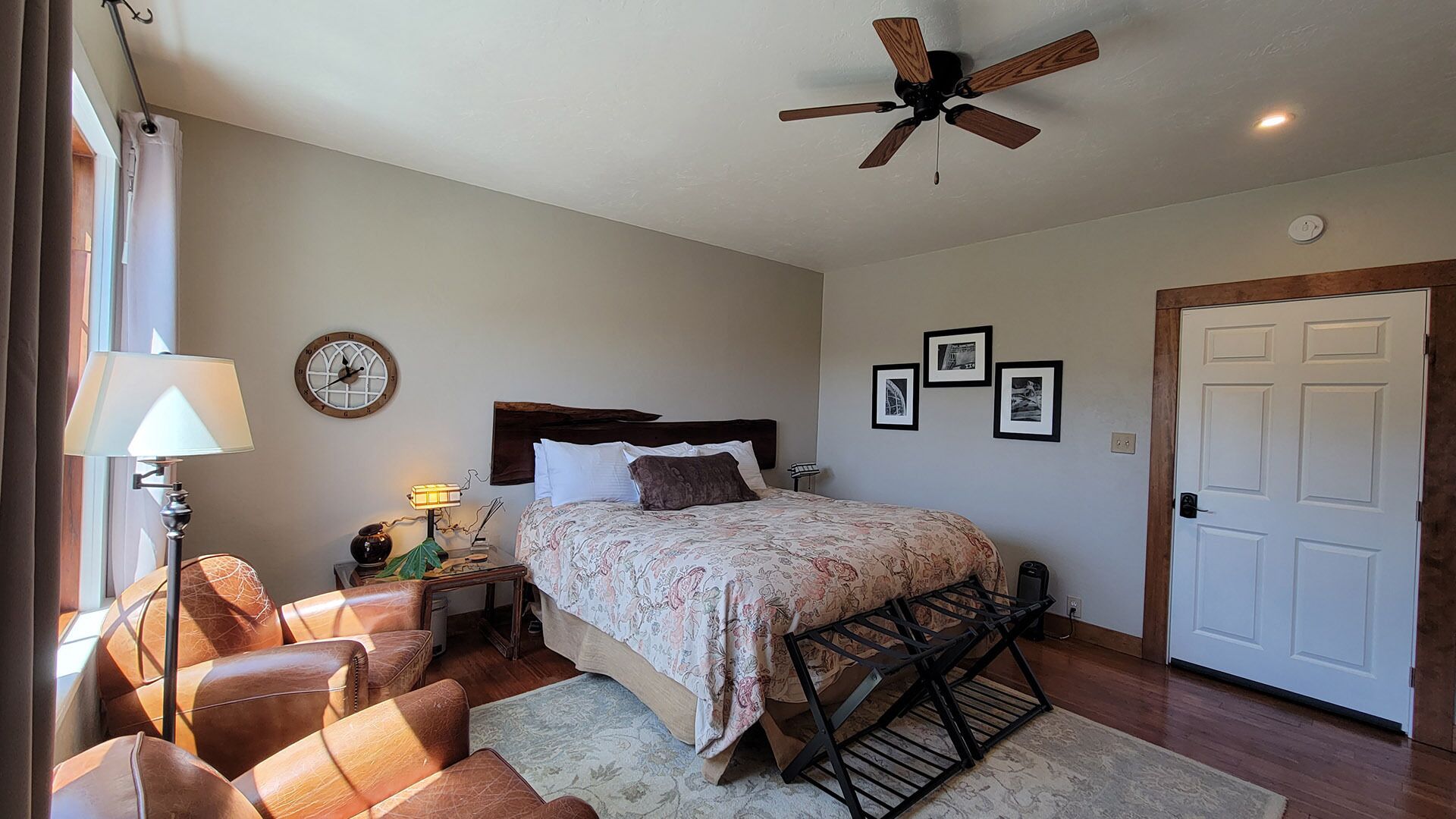 Une chambre à coucher comprenant un lit, une table de chevet, une lampe, une chaise, un ventilateur de plafond et une porte.